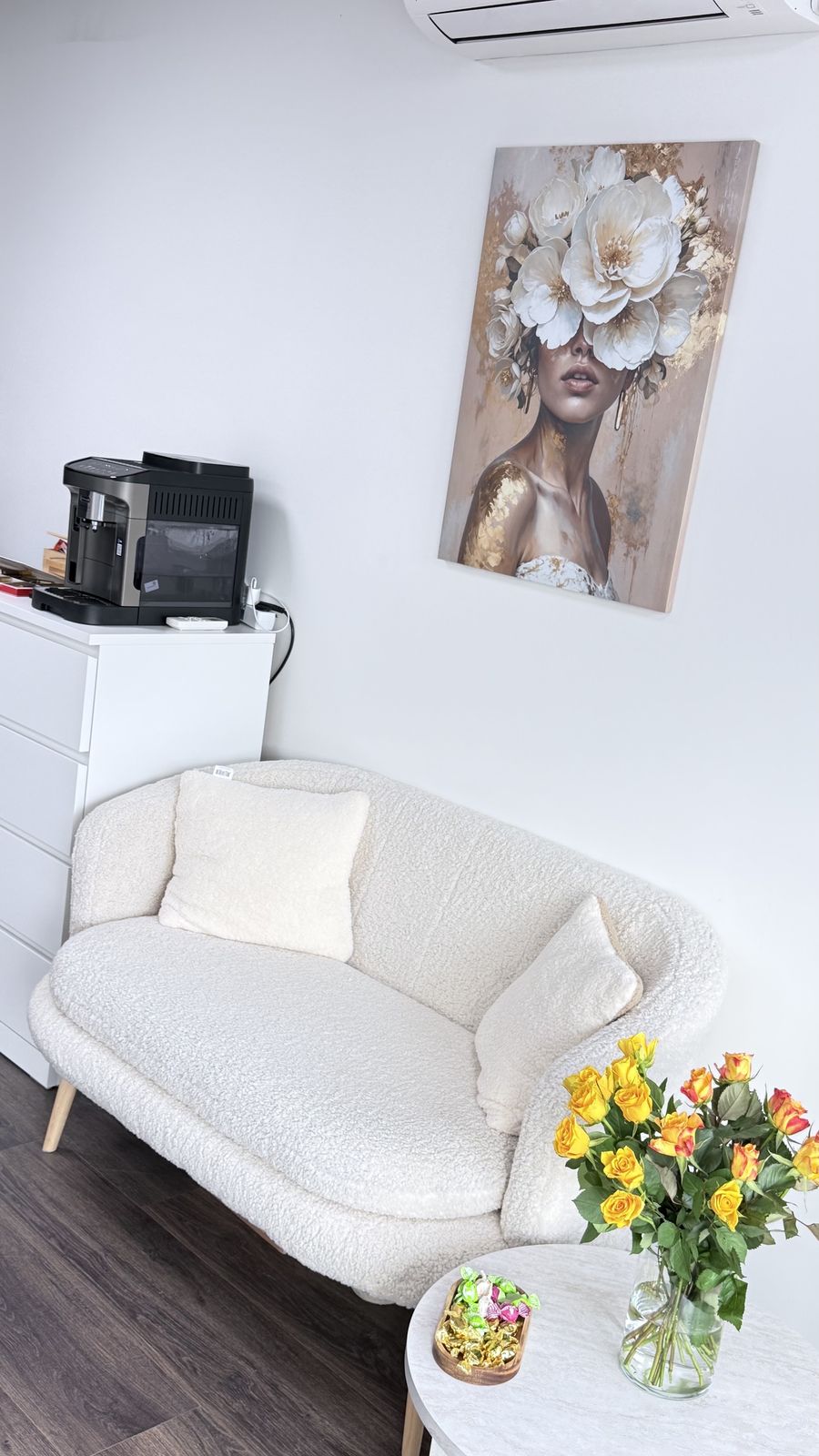 Een witte bank, een koffieapparaat en een schilderij van een vrouw met bloemen